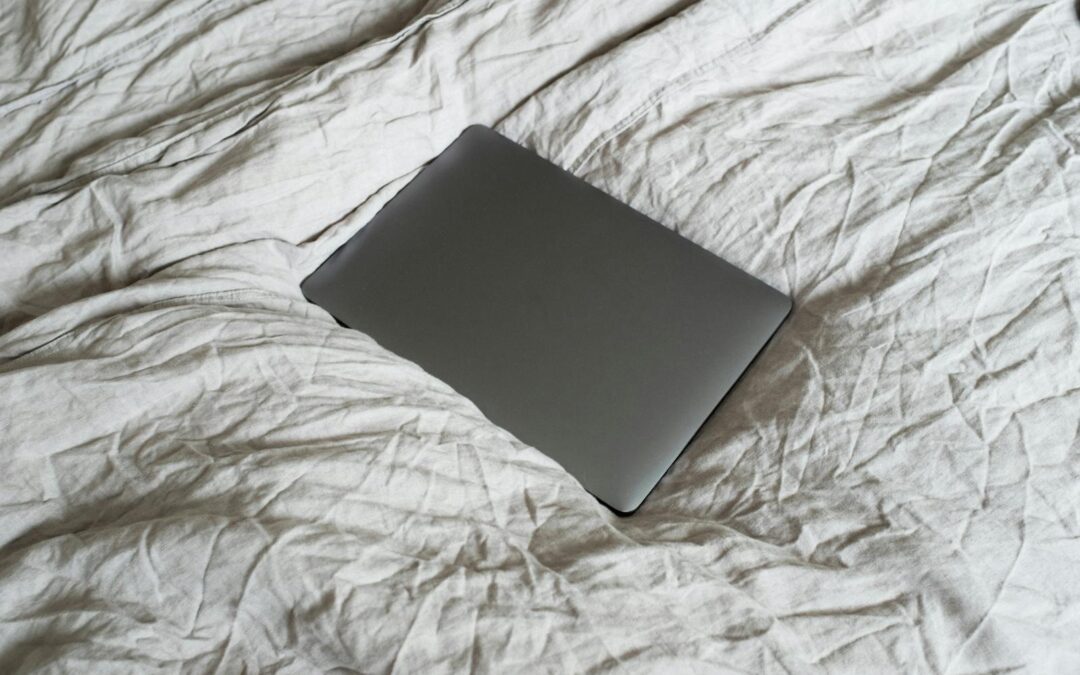 From above of closed dark gray portable computer located on large soft and crumpled cotton bed sheet in apartment in light room